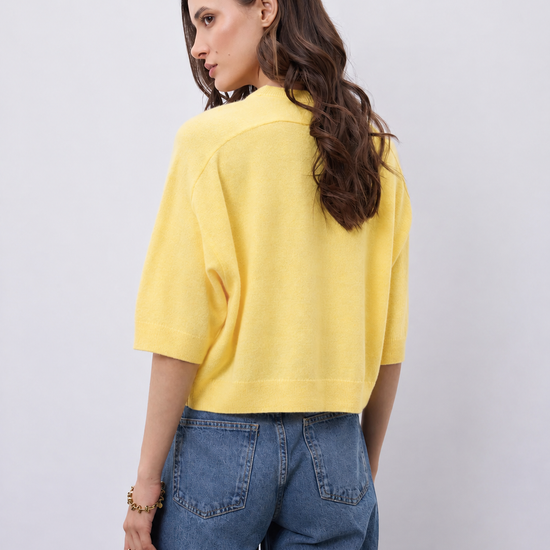 A woman shown from the back wearing a bright yellow knit short-sleeve crop top and blue jeans, standing against a light gray studio background.