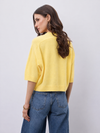 A woman shown from the back wearing a bright yellow knit short-sleeve crop top and blue jeans, standing against a light gray studio background.