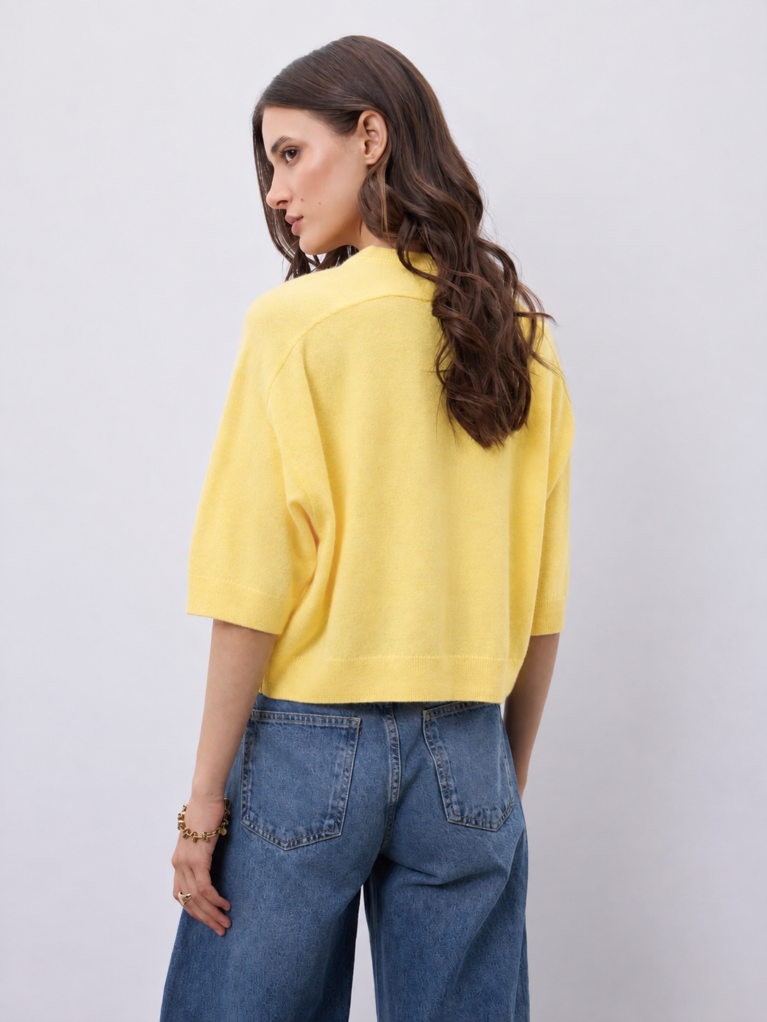 A woman shown from the back wearing a bright yellow knit short-sleeve crop top and blue jeans, standing against a light gray studio background.