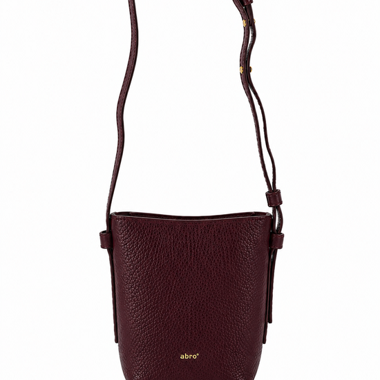 A dark burgundy pebbled leather crossbody bag with a thin matching leather strap and a small gold logo on the front, shown against a plain white background.