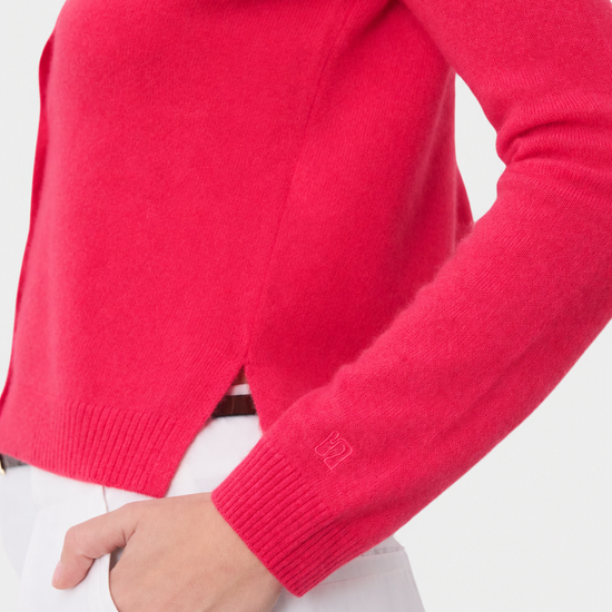 A close-up of a person wearing a bright pink knit sweater with white trousers, showing the ribbed cuffs and a small tonal logo patch on the sleeve.