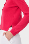 A close-up of a person wearing a bright pink knit sweater with white trousers, showing the ribbed cuffs and a small tonal logo patch on the sleeve.
