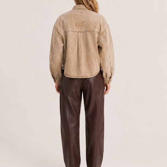 A model viewed from the back wearing a cropped beige suede jacket with a tonal embroidered logo on the upper back, paired with dark brown wide-leg trousers and black shoes, against a light beige studio background.