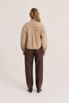 A model viewed from the back wearing a cropped beige suede jacket with a tonal embroidered logo on the upper back, paired with dark brown wide-leg trousers and black shoes, against a light beige studio background.