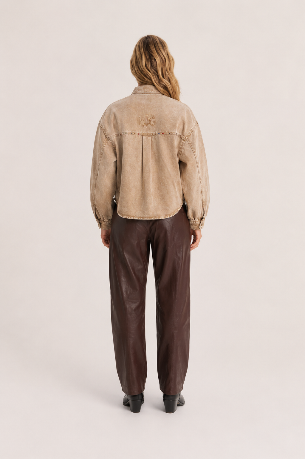 A model viewed from the back wearing a cropped beige suede jacket with a tonal embroidered logo on the upper back, paired with dark brown wide-leg trousers and black shoes, against a light beige studio background.