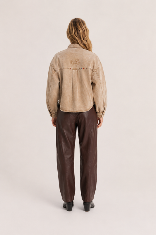 A model viewed from the back wearing a cropped beige suede jacket with a tonal embroidered logo on the upper back, paired with dark brown wide-leg trousers and black shoes, against a light beige studio background.
