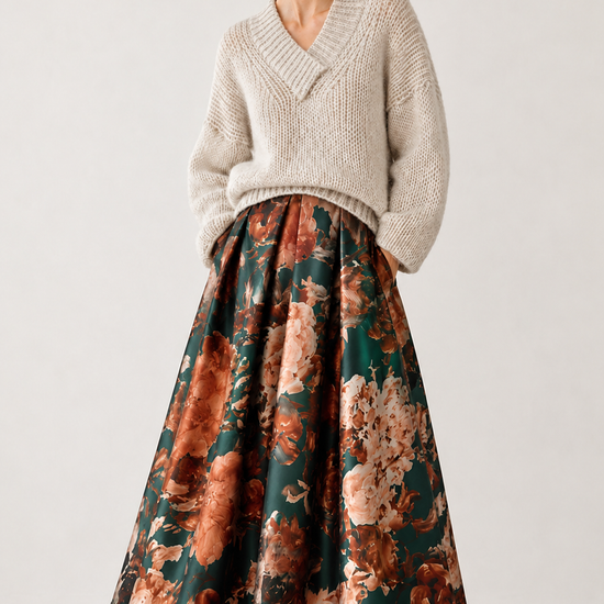 A model wearing a cream-colored knit V-neck sweater and a long, flared skirt with a brown, orange, and green floral pattern, paired with black pointed-toe heels, set against a plain light gray studio background.