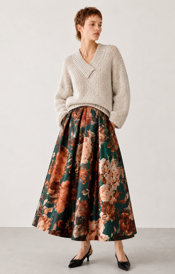 A model wearing a cream-colored knit V-neck sweater and a long, flared skirt with a brown, orange, and green floral pattern, paired with black pointed-toe heels, set against a plain light gray studio background.