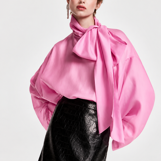 A model wearing a vibrant pink satin blouse with dramatic balloon sleeves and a large bow at the neck, paired with a black textured faux-leather skirt, against a light gray studio background.