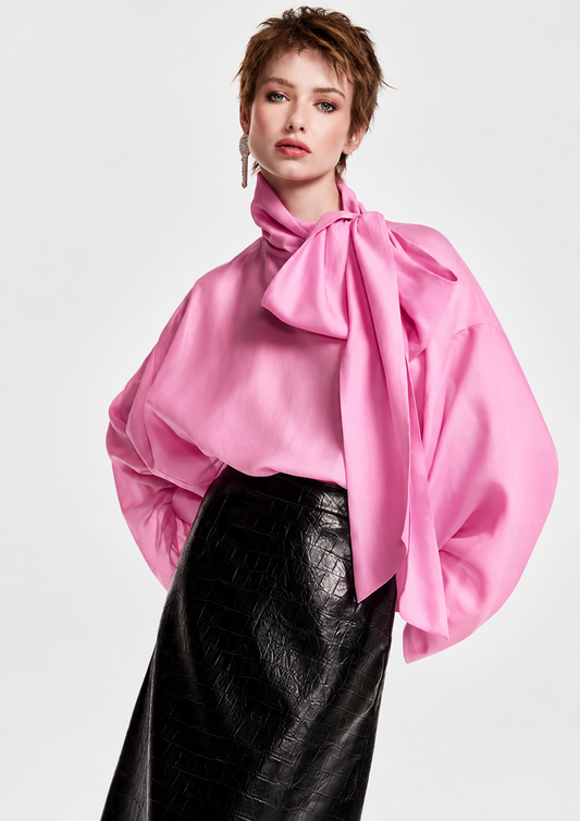 A model wearing a vibrant pink satin blouse with dramatic balloon sleeves and a large bow at the neck, paired with a black textured faux-leather skirt, against a light gray studio background.
