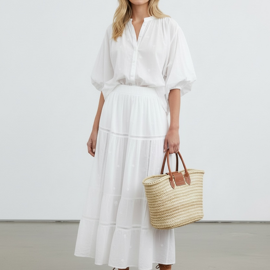 Woman in a white outfit holding a straw bag against a plain background