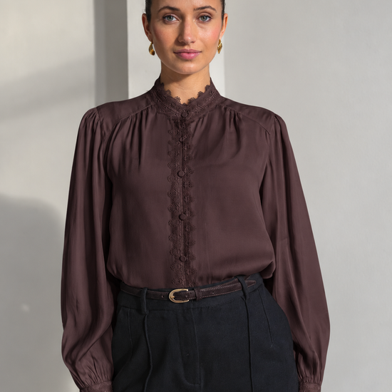 A woman modeling a dark brown long-sleeve blouse with a lace-trimmed high neck and button placket, paired with black trousers, standing against a light gray wall.