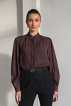 A woman modeling a dark brown long-sleeve blouse with a lace-trimmed high neck and button placket, paired with black trousers, standing against a light gray wall.