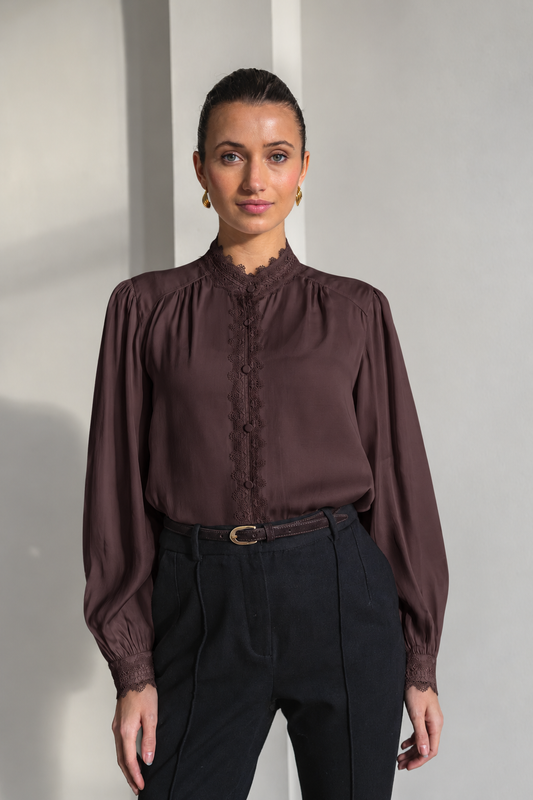 A woman modeling a dark brown long-sleeve blouse with a lace-trimmed high neck and button placket, paired with black trousers, standing against a light gray wall.
