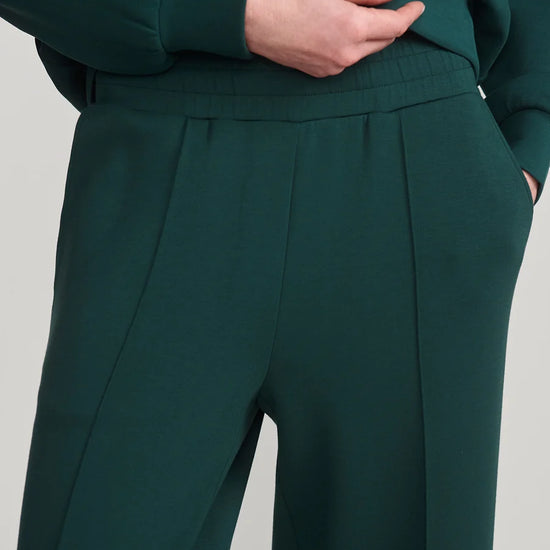 Close-up of a person wearing dark green wide-leg trousers with a matching top, showing the waistband, front pleats, and hand in pocket against a plain light gray background.