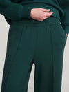 Close-up of a person wearing dark green wide-leg trousers with a matching top, showing the waistband, front pleats, and hand in pocket against a plain light gray background.