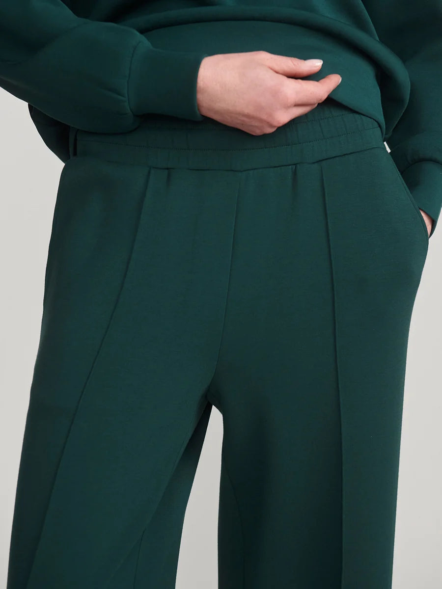 Close-up of a person wearing dark green wide-leg trousers with a matching top, showing the waistband, front pleats, and hand in pocket against a plain light gray background.