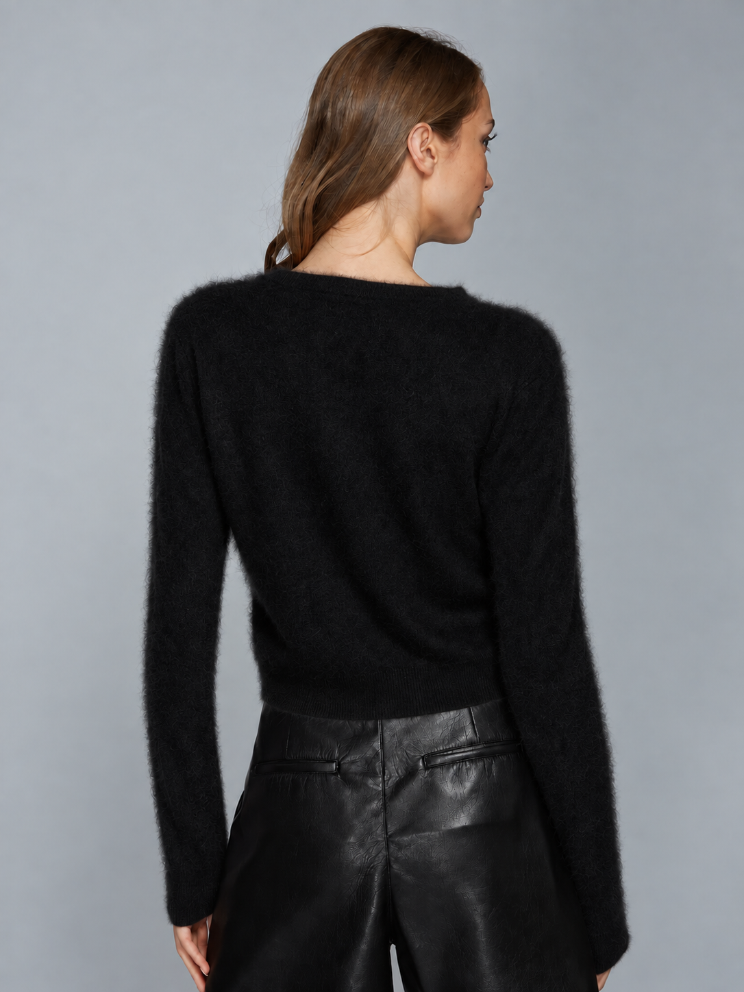 A woman shown from the back wearing a black fuzzy long-sleeve sweater and black leather pants against a plain gray studio background.