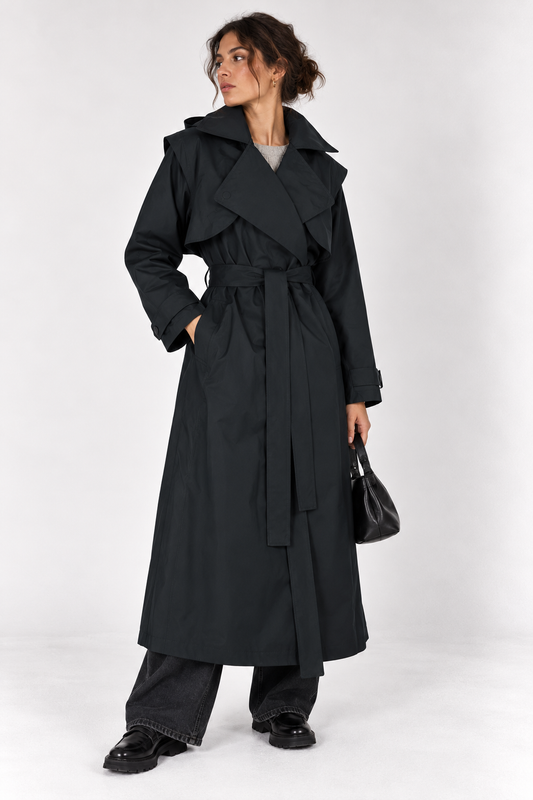 model shown wearing a long black trench coat with her hand in a pocket whikst holding a bag on the other hand and standing on a plain white background. 