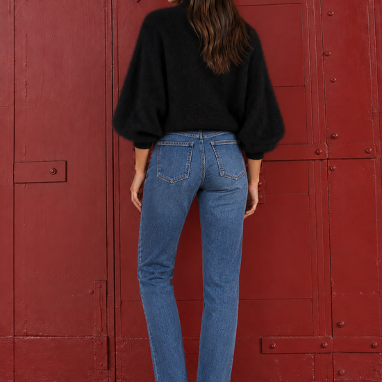 A woman is shown from behind wearing a black long-sleeve knit sweater and blue jeans, standing in front of a large red metal door.