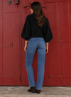 A woman is shown from behind wearing a black long-sleeve knit sweater and blue jeans, standing in front of a large red metal door.