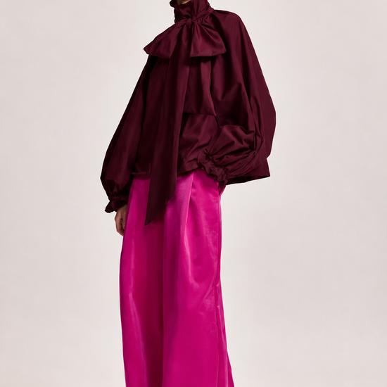 A model wearing a dark burgundy high-neck blouse with dramatic ruffles and voluminous sleeves, paired with bright fuchsia wide-leg trousers, against a plain light gray studio background.