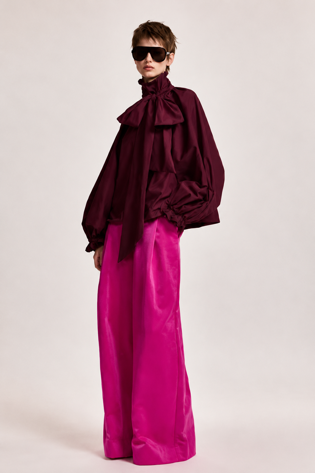 A model wearing a dark burgundy high-neck blouse with dramatic ruffles and voluminous sleeves, paired with bright fuchsia wide-leg trousers, against a plain light gray studio background.