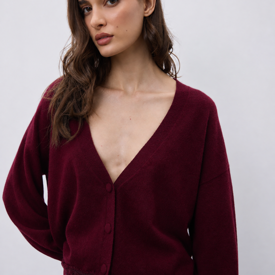 A woman wearing a deep burgundy cardigan with a V-neck, button closure, and lace trim at the hem, paired with blue jeans against a plain light gray background.