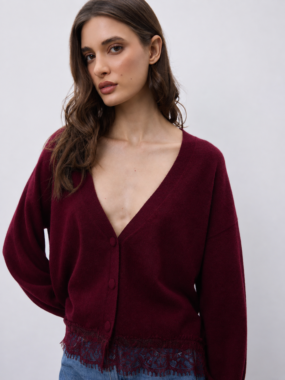A woman wearing a deep burgundy cardigan with a V-neck, button closure, and lace trim at the hem, paired with blue jeans against a plain light gray background.