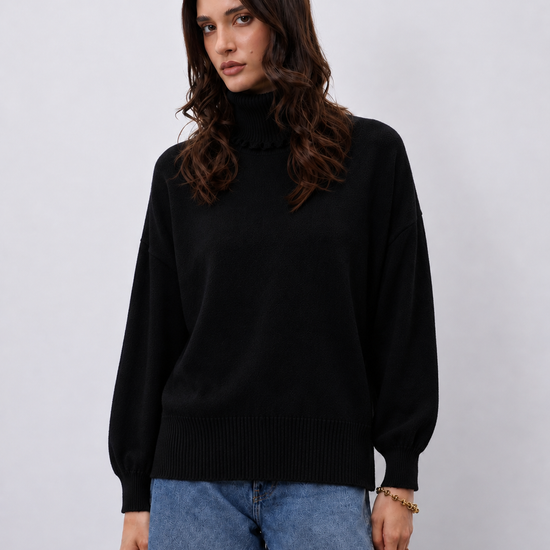 A woman wearing a black ribbed mock-neck sweater and blue jeans, standing against a light gray studio background.