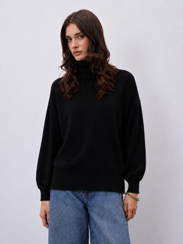 A woman wearing a black ribbed mock-neck sweater and blue jeans, standing against a light gray studio background.