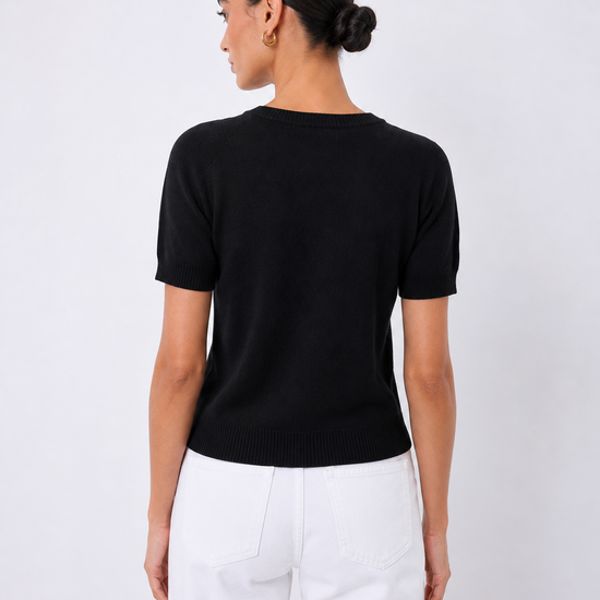 A woman shown from the back wearing a black short-sleeve knit top and white pants against a light gray studio background.