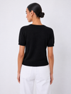 A woman shown from the back wearing a black short-sleeve knit top and white pants against a light gray studio background.