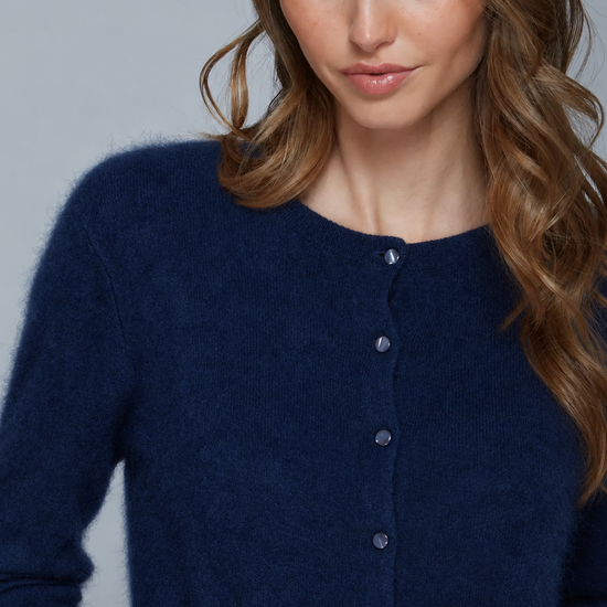A close-up of a woman wearing a fuzzy navy blue button-front cardigan sweater against a plain light gray studio background.