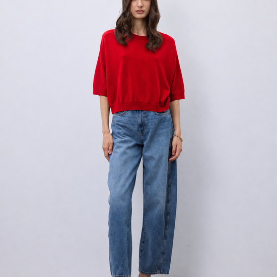 A woman stands against a light gray studio wall wearing a bright red short-sleeve top, blue straight-leg jeans, and beige snakeskin heels.