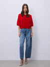 A woman stands against a light gray studio wall wearing a bright red short-sleeve top, blue straight-leg jeans, and beige snakeskin heels.