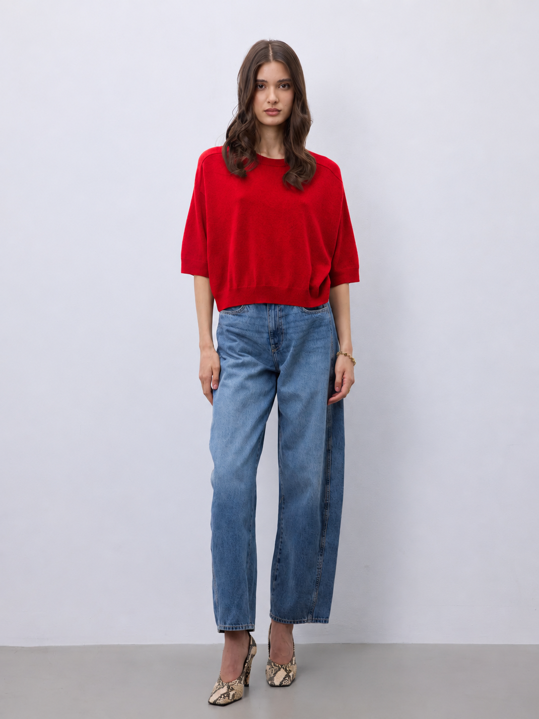 A woman stands against a light gray studio wall wearing a bright red short-sleeve top, blue straight-leg jeans, and beige snakeskin heels.