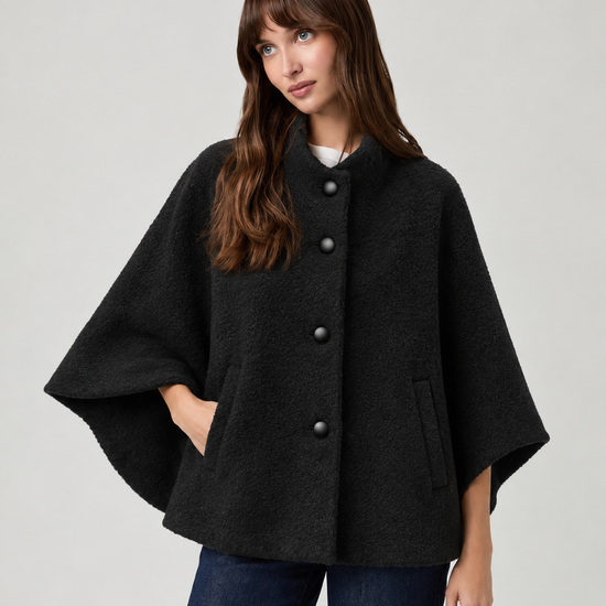 model wearing black cape style coat with blue denim jeans and black boots on a plain white background