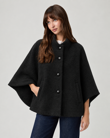 model wearing black cape style coat with blue denim jeans and black boots on a plain white background