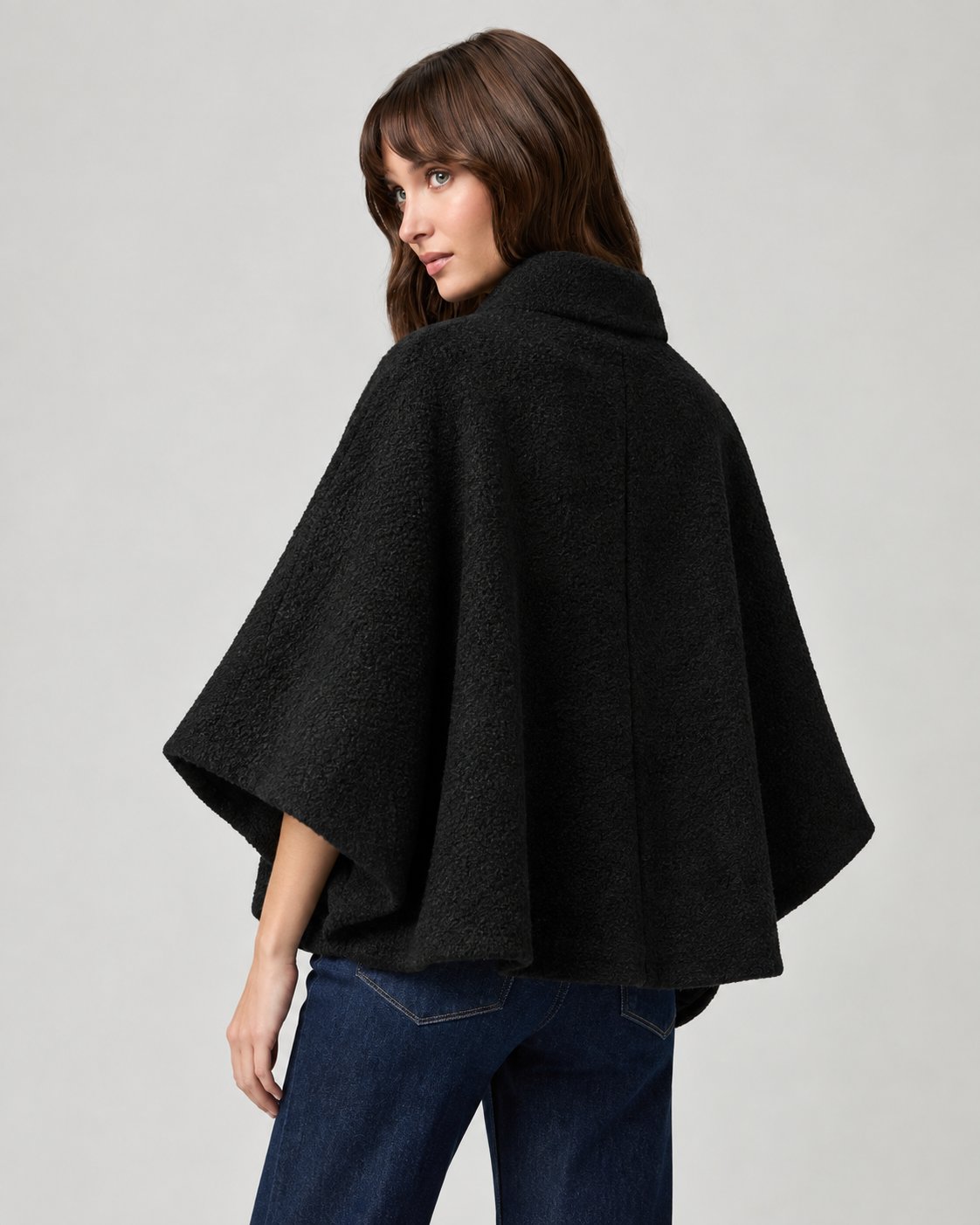 model wearing black cape style coat with blue denim jeans and black boots on a plain white background