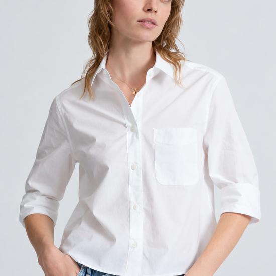 A woman wearing a white long-sleeve button-up shirt with a chest pocket, paired with light blue jeans, against a light gray studio background.