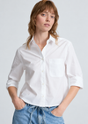 A woman wearing a white long-sleeve button-up shirt with a chest pocket, paired with light blue jeans, against a light gray studio background.