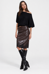 A model wearing a black off-the-shoulder knit top, a shiny dark brown faux-leather midi skirt, and black knee-high boots, posed against a light gray studio background.