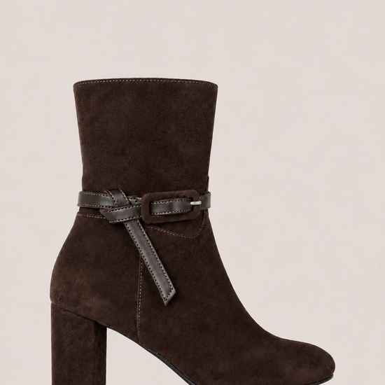 A dark brown suede ankle boot with a block heel and a matching dark brown leather strap detail around the ankle, shown against a solid light beige background.
