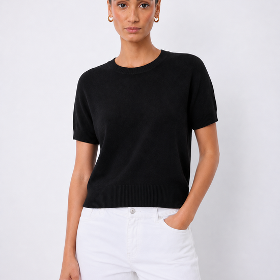A woman modeling a short-sleeve black knit crewneck sweater paired with white pants, standing against a light gray studio background.