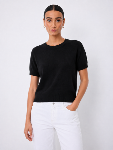 A woman modeling a short-sleeve black knit crewneck sweater paired with white pants, standing against a light gray studio background.