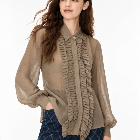 A woman modeling a taupe sheer long-sleeve blouse with a ruffled front panel and button collar, paired with dark blue patterned pants, against a plain white studio background.
