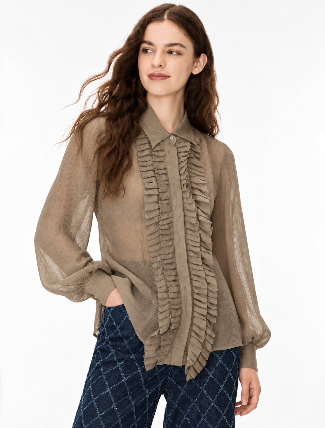 A woman modeling a taupe sheer long-sleeve blouse with a ruffled front panel and button collar, paired with dark blue patterned pants, against a plain white studio background.