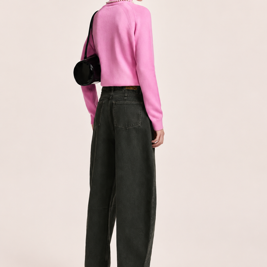 A model is shown from the back wearing a bright pink long-sleeve knit sweater, dark charcoal gray wide-leg jeans, black ankle boots, a black baseball cap, and carrying a black round shoulder bag against a solid light beige background.