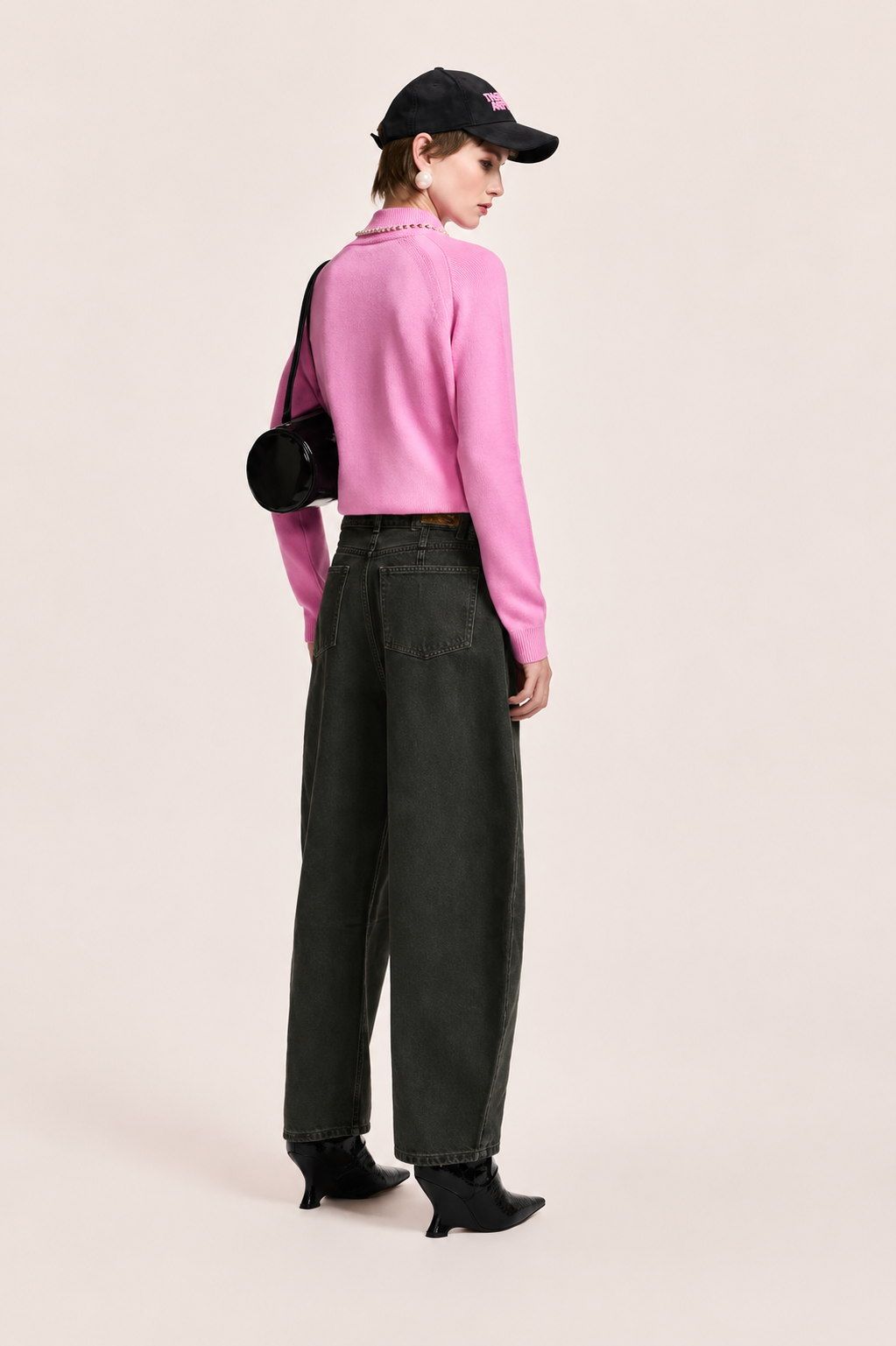 A model is shown from the back wearing a bright pink long-sleeve knit sweater, dark charcoal gray wide-leg jeans, black ankle boots, a black baseball cap, and carrying a black round shoulder bag against a solid light beige background.